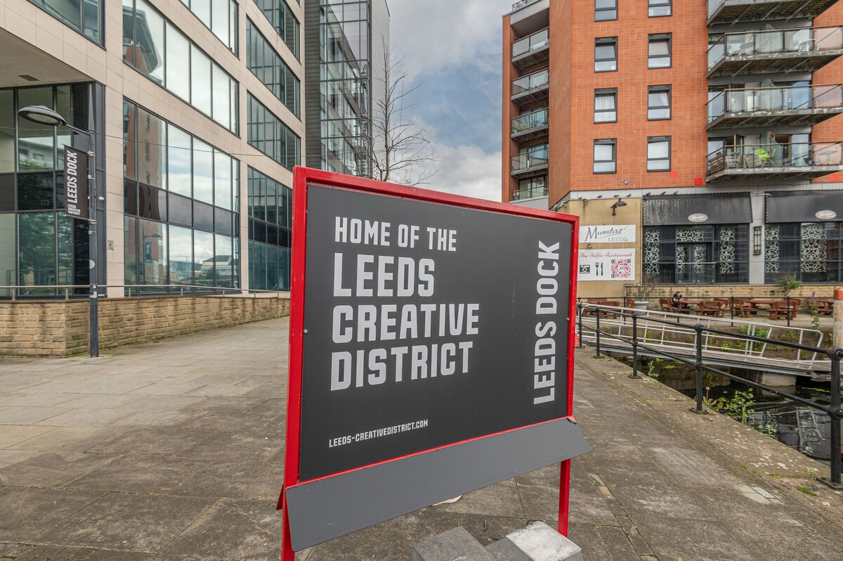 A large sign denotes the Leeds Creative District, set against a backdrop of modern buildings and a waterway. The area features a mix of glass and red-brick structures, indicating an urban environment with easy access to arts and culture.