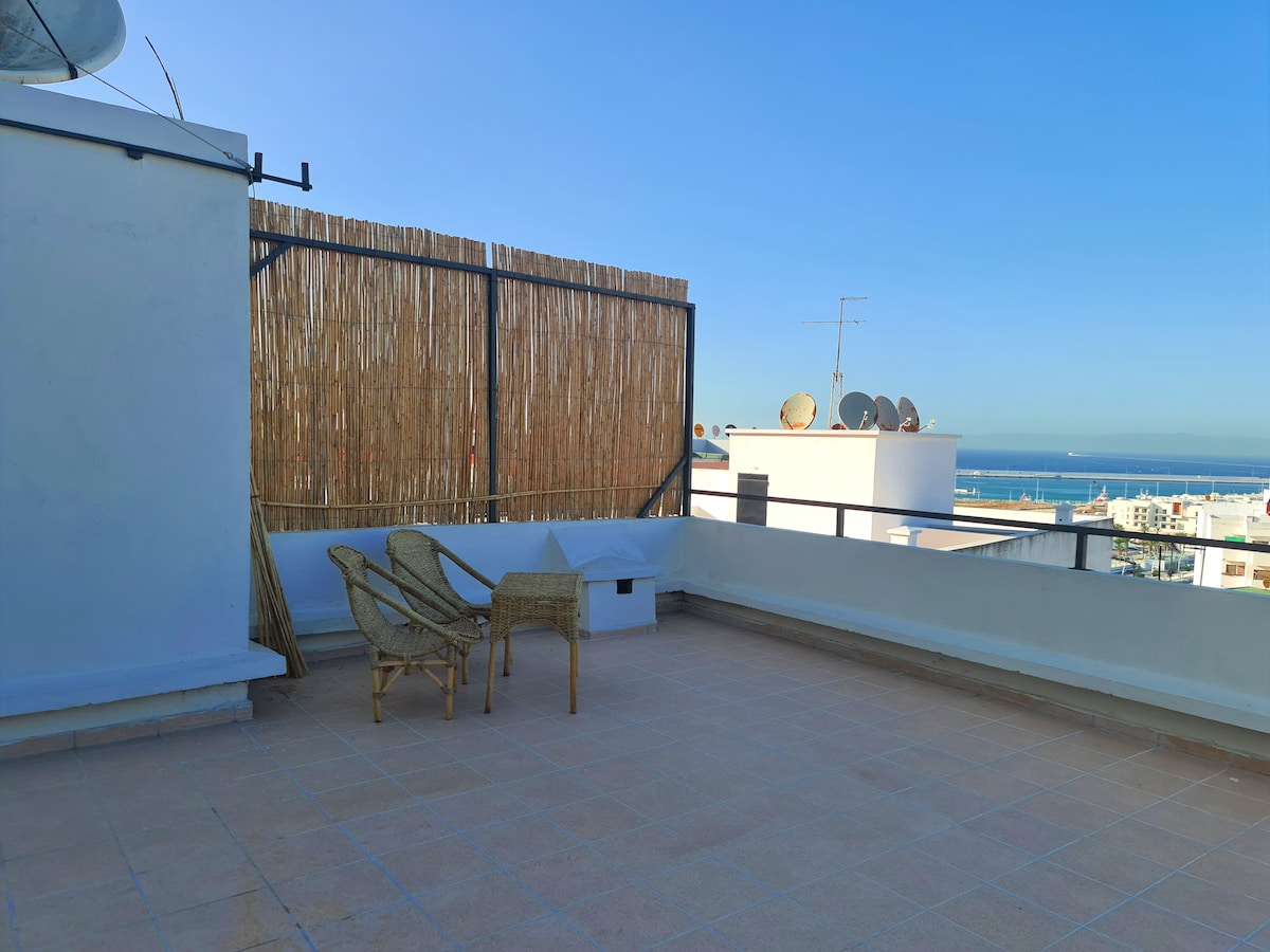 A private terrace with a panoramic view of the sea and the Strait of Gibraltar is depicted. Two woven chairs and a small table are situated on the tiled floor, surrounded by a wooden privacy screen. The sky is clear and blue, enhancing the serene atmosphere.