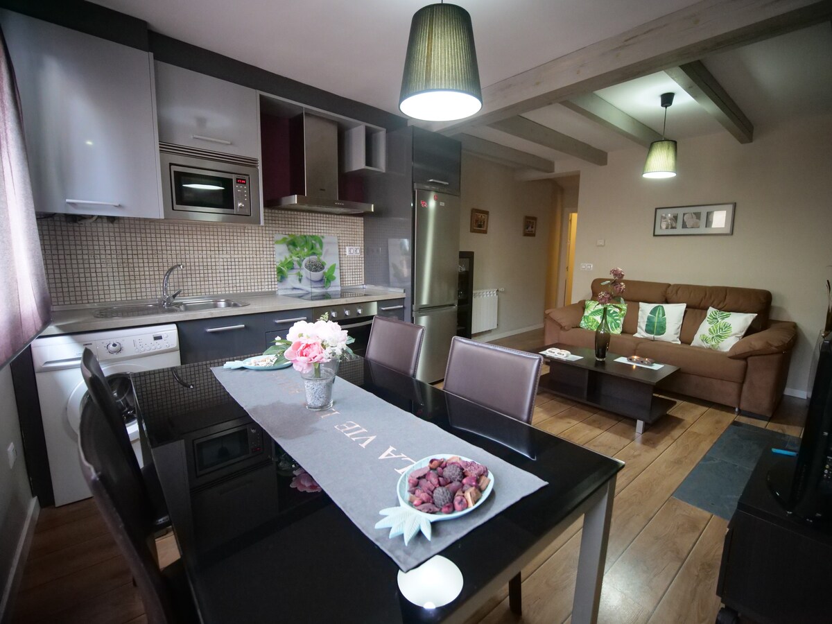 A modern kitchen and dining area feature sleek cabinetry and a large dining table. A comfortable sofa is positioned in the living space, complemented by decorative cushions. Natural light enters through a window, enhancing the space's welcoming ambiance.