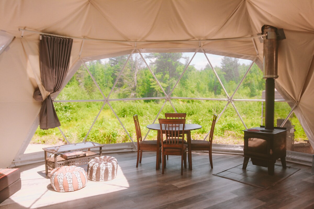 The interior of the geodesic dome features a round wooden table with four chairs positioned near a cozy wood stove. Two poufs are placed on the floor, while large windows provide a view of the surrounding greenery, enhancing the natural light within the space.