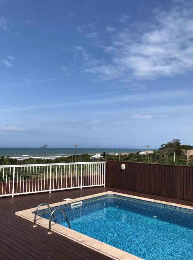 A private pool is surrounded by a wooden deck, providing clear views of the ocean and a bright blue sky. The nearby beach is visible, complemented by distant seaside lights and greenery in the background.