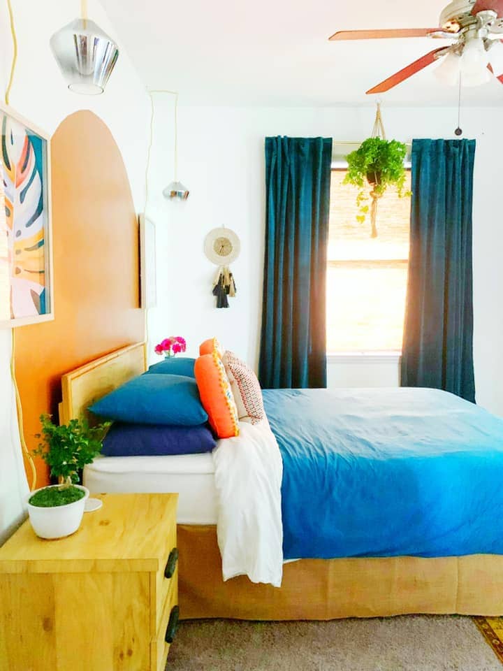 A bright bedroom features a spacious bed with a blue coverlet, accented by vibrant orange and patterned pillows. Dark curtains frame a window, allowing natural light to illuminate the space. A potted plant enhances the wooden nightstand, while wall art adds a touch of color.