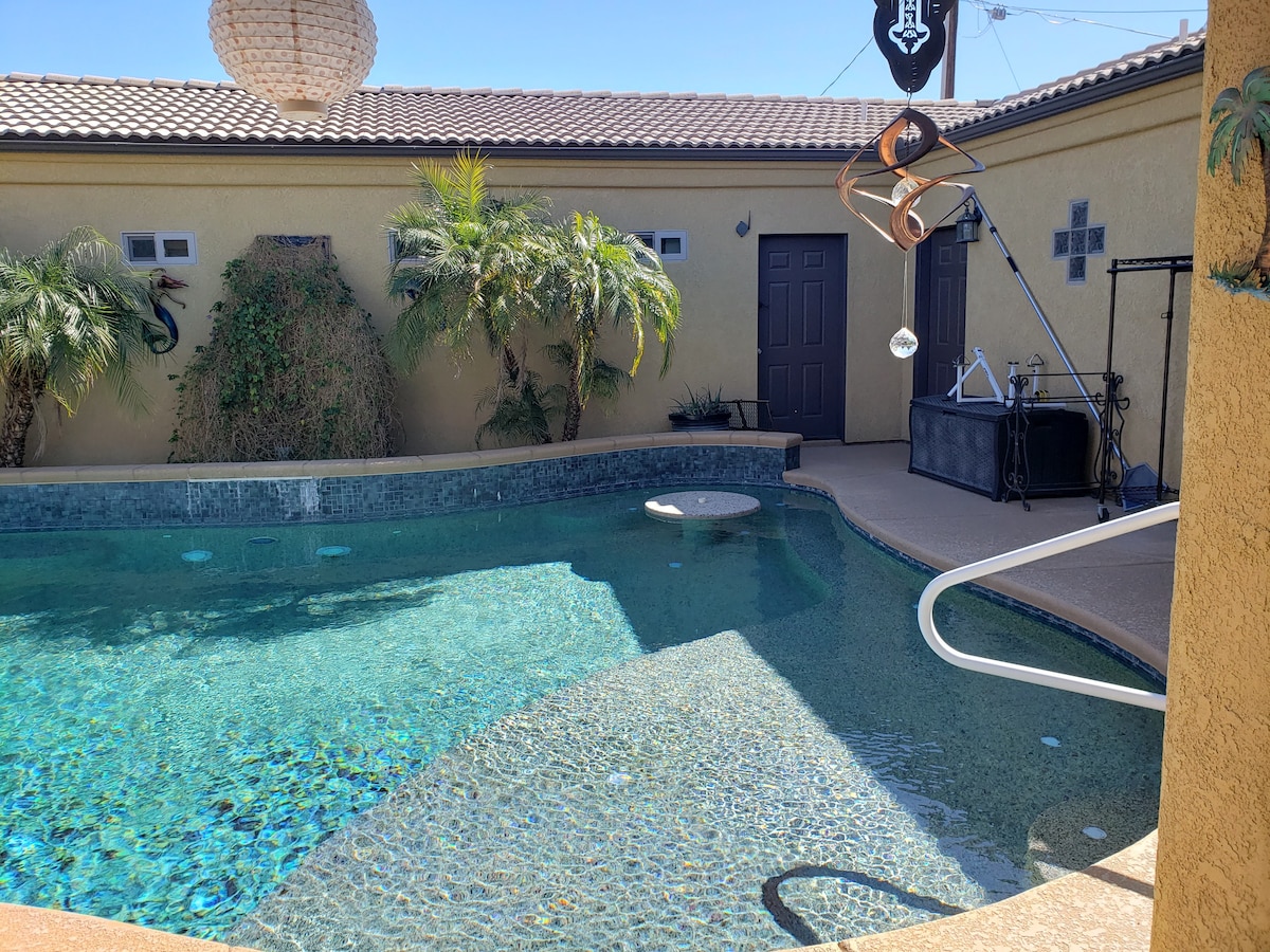 A private pool area is framed by tropical plants and a textured wall, featuring clear blue water that reflects sunlight. A sunlit patio provides space for relaxation, while a nearby structure offers easy access to the outdoor amenities. The setting is designed for tranquility.