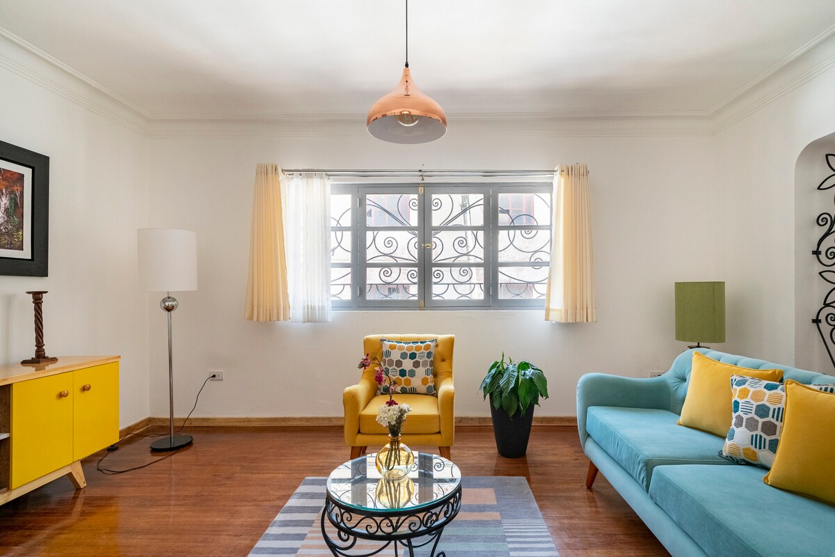 A bright living area features a blue sofa and a vibrant yellow armchair, accented by decorative cushions. A round glass coffee table is positioned at the center. Natural light enters through large windows with sheer curtains, complemented by pastel yellow drapes.