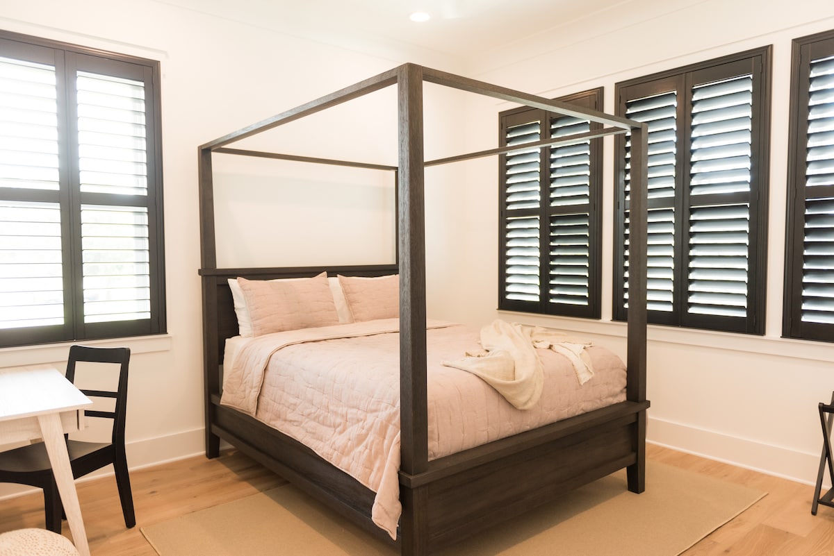 A four-poster bed with a soft pink quilt is positioned centrally in the room, surrounded by shuttered windows. A small table and chair set is located to the side, with light-colored flooring adding to the serene atmosphere.