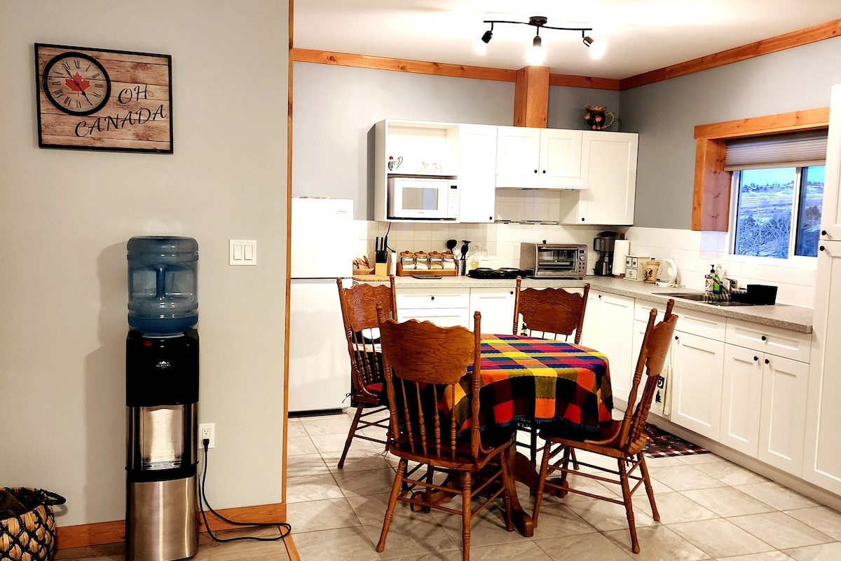 The kitchen area features a wooden dining table surrounded by six chairs, covered with a colorful tablecloth. Appliances, including a microwave and coffee maker, are visible on the countertops. A water cooler stands nearby, and a window allows natural light to illuminate the space.