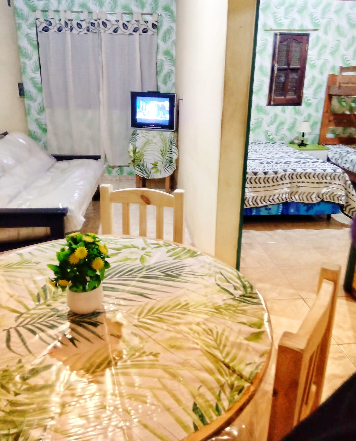 A living space features a round table with a plant centerpiece, surrounded by wooden chairs. A compact television is visible beside a cozy seating area, with a spacious bed positioned in the background, adorned with a striped blanket. Green patterned walls create a fresh ambiance.