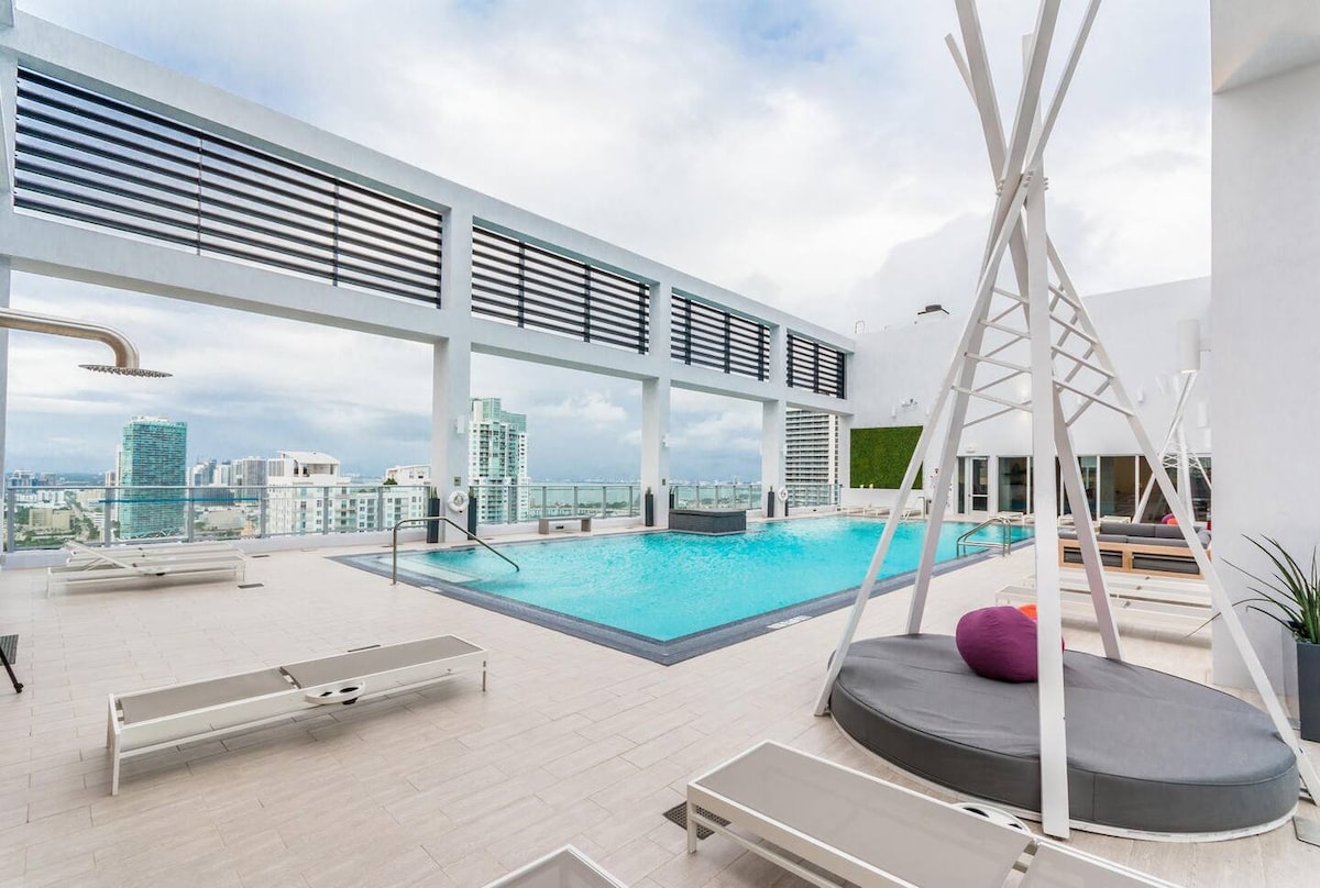 A spacious rooftop pool area is highlighted by a large swimming pool surrounded by sun loungers and shaded seating. The skyline of the city is visible in the background, providing a scenic view. Natural light fills the space through the large, open design.
