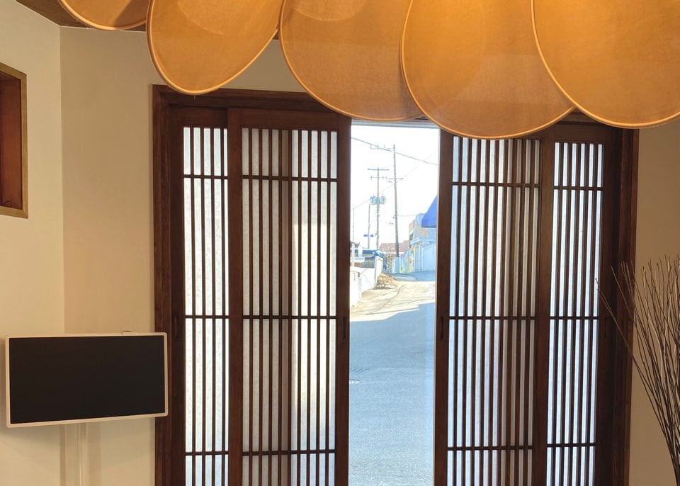 Sliding wooden doors with a traditional lattice design are seen, allowing natural light to filter into the room. Above, a series of curved light fixtures hang, creating a gentle ambiance. A flat-screen television is positioned on a stand nearby, complementing the space.