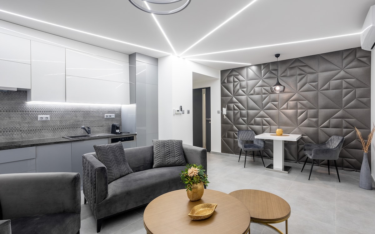 A modern living area features a geometric accent wall and sleek furniture. A comfortable sofa and two chairs surround a coffee table, while a dining table is positioned nearby. Bright lighting highlights the contemporary design and the kitchen area is visible, showcasing a minimalist aesthetic.