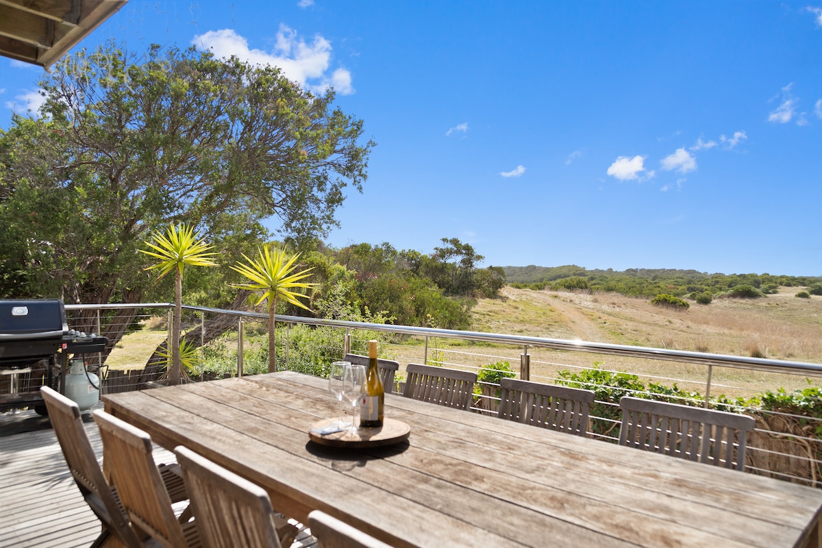 A spacious outdoor deck features a large wooden dining table surrounded by multiple chairs. A barbecue is positioned nearby, and the scenic countryside offers views of rolling hills and greenery under a clear blue sky.
