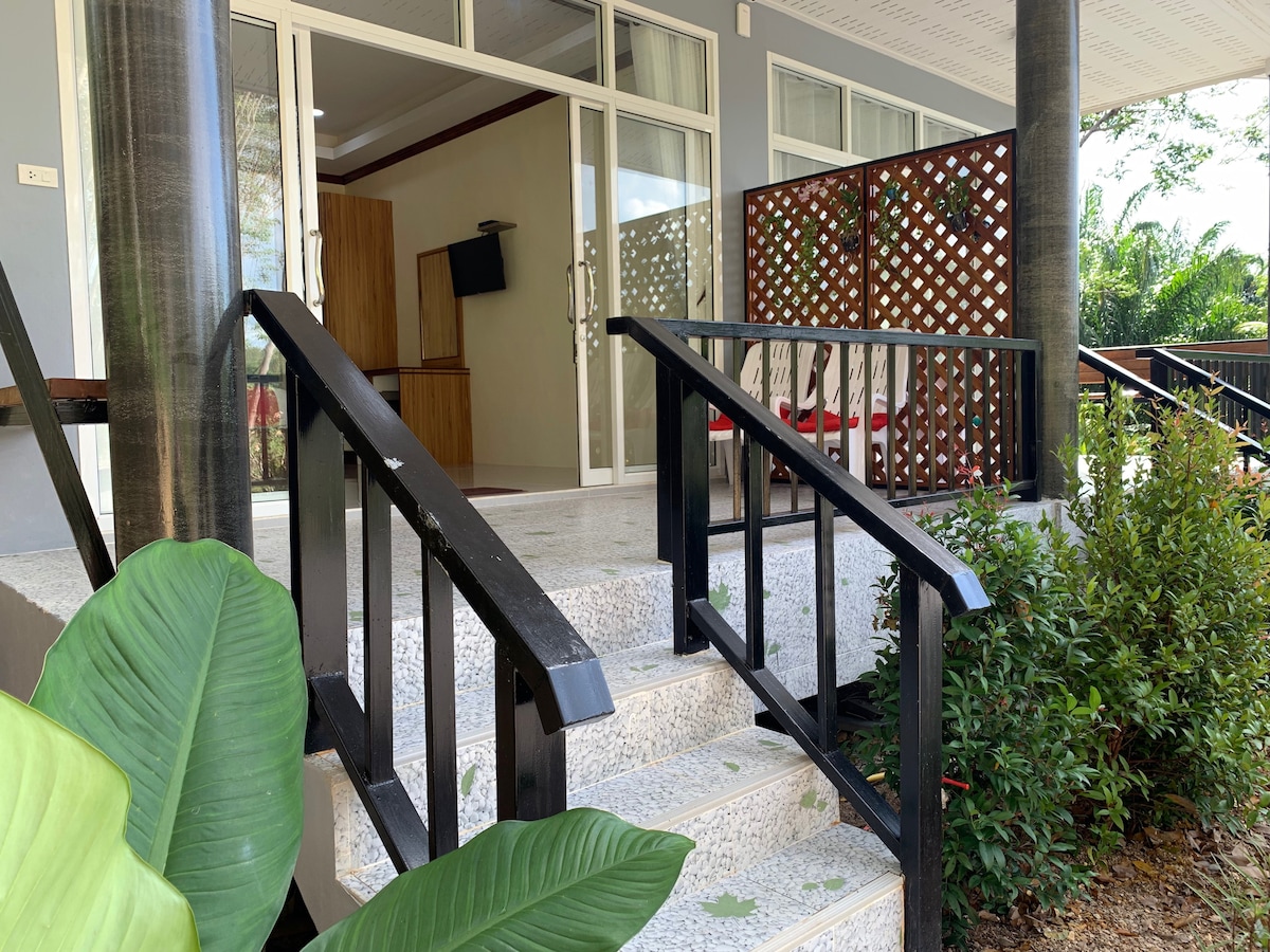The entrance features a set of steps leading to a covered patio area. A modern design with black railings and white stone steps enhances accessibility. Lush green foliage and a decorative wooden screen provide privacy, while a glimpse of the interior room with a television is visible inside.