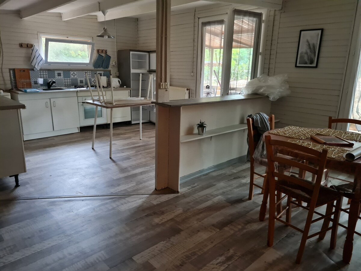 An open-concept kitchen and dining area is visible, featuring a modern design with wood flooring. The kitchen contains a refrigerator, stove, and ample counter space, while a dining table with wooden chairs is situated nearby. Large windows allow natural light to fill the space.