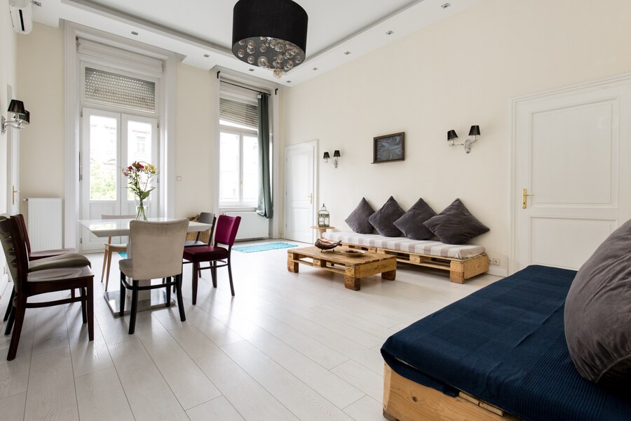 A spacious living area features a dining table surrounded by chairs, complemented by a cozy seating arrangement with floor cushions. Natural light floods through large windows, highlighting the light-colored wood flooring and minimalistic decor.