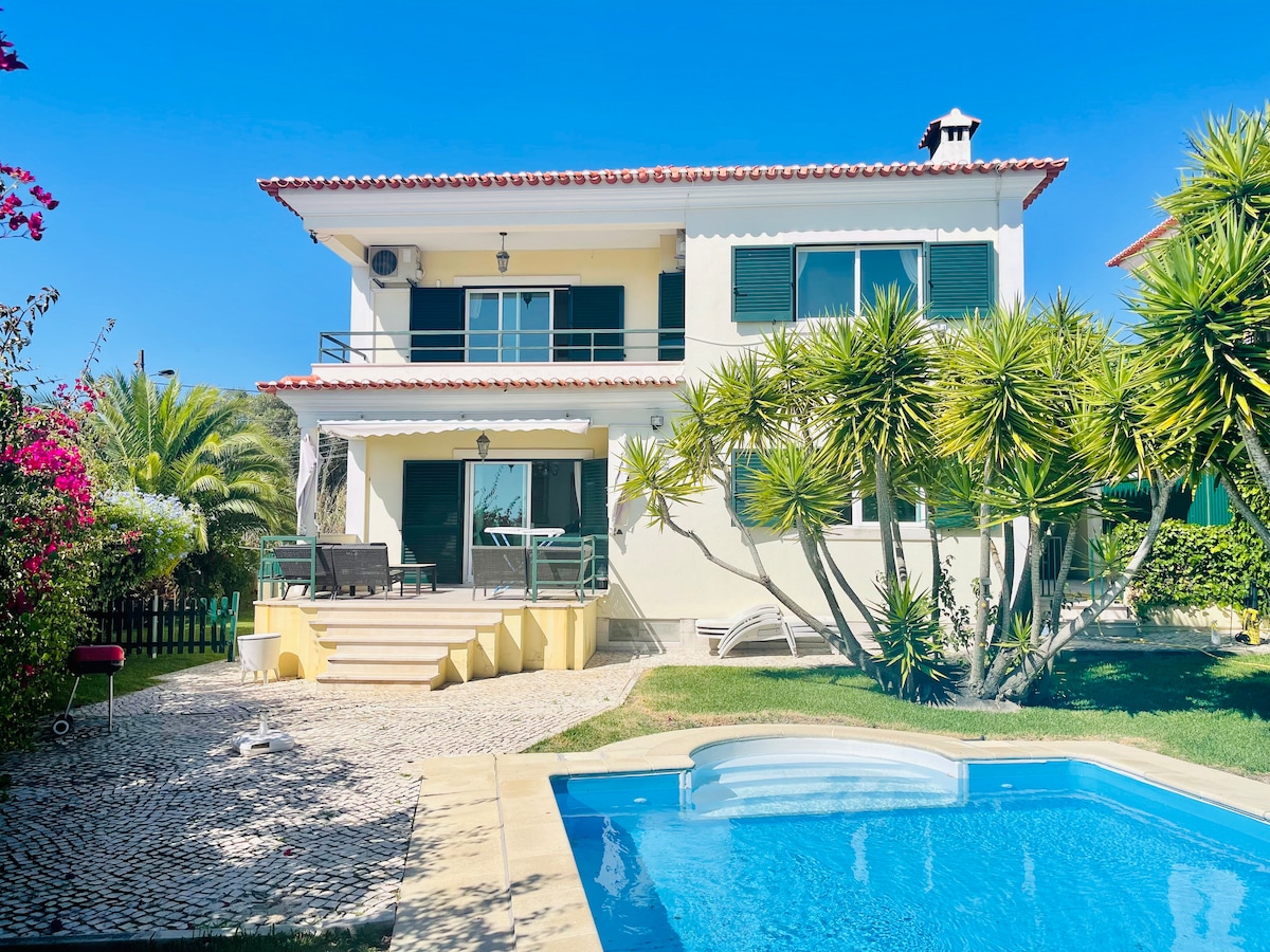 The two-story villa features a large garden area with tropical plants and palm trees. A crystal-clear pool is visible in the foreground, while a spacious patio with seating is situated in front of the house, which has large windows and a tile roof.