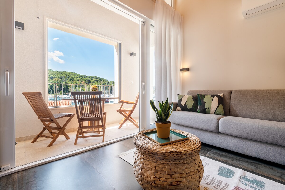 A bright living area features a comfortable gray sofa paired with a natural wicker coffee table. Two wooden chairs are positioned on the balcony, offering views of the greenery outside. Large sliding glass doors open to create a seamless connection with the outdoors.