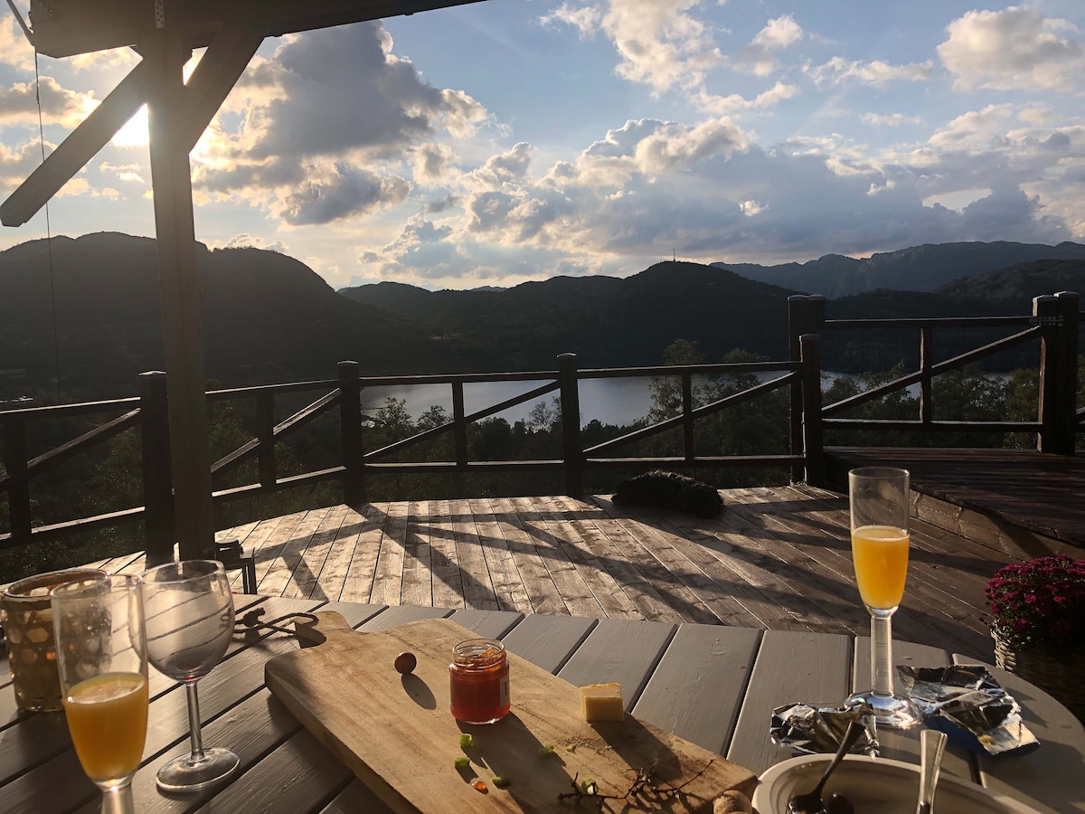 A spacious wooden deck is shown overlooking a scenic landscape of mountains and water. A charcuterie board sits prominently, accompanied by a glass of sparkling beverage and a cocktail. The sky is partly cloudy, casting soft light over the surrounding nature.