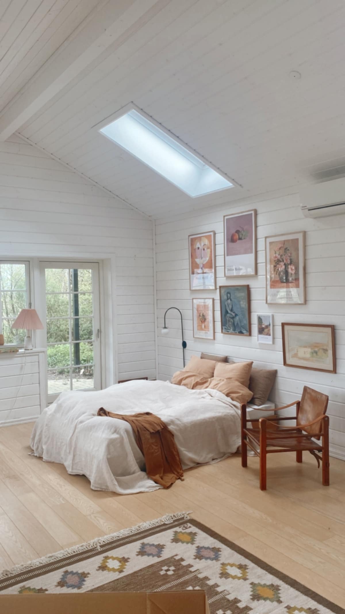 An open room features a spacious bed with soft bedding and a cozy throw. Art pieces adorn the white walls, and natural light enters through a skylight and glass door, leading to a view of a garden. A wooden chair complements the inviting space.
