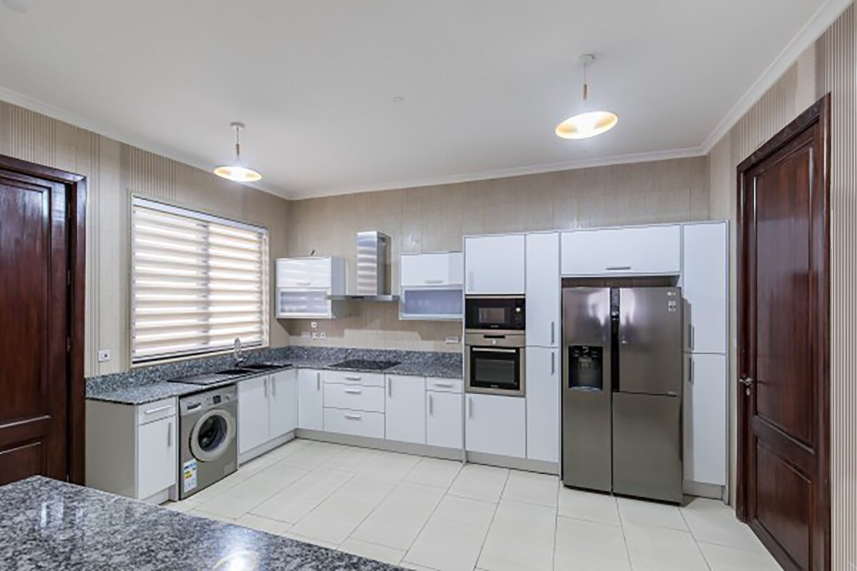 A modern kitchen is featured, showcasing white cabinetry and a dark granite countertop. Stainless steel appliances are included, such as a refrigerator and an oven. A washing machine is positioned beside the kitchen area, with ample natural light filtering through layered window treatments.