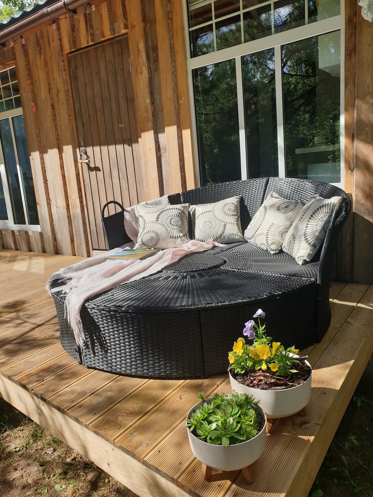A circular outdoor seating area is presented on a wooden deck, featuring dark wicker furniture adorned with decorative pillows. Nearby, potted plants with colorful blooms add a touch of greenery, complementing the natural setting of the wooden cabin and surrounding trees.