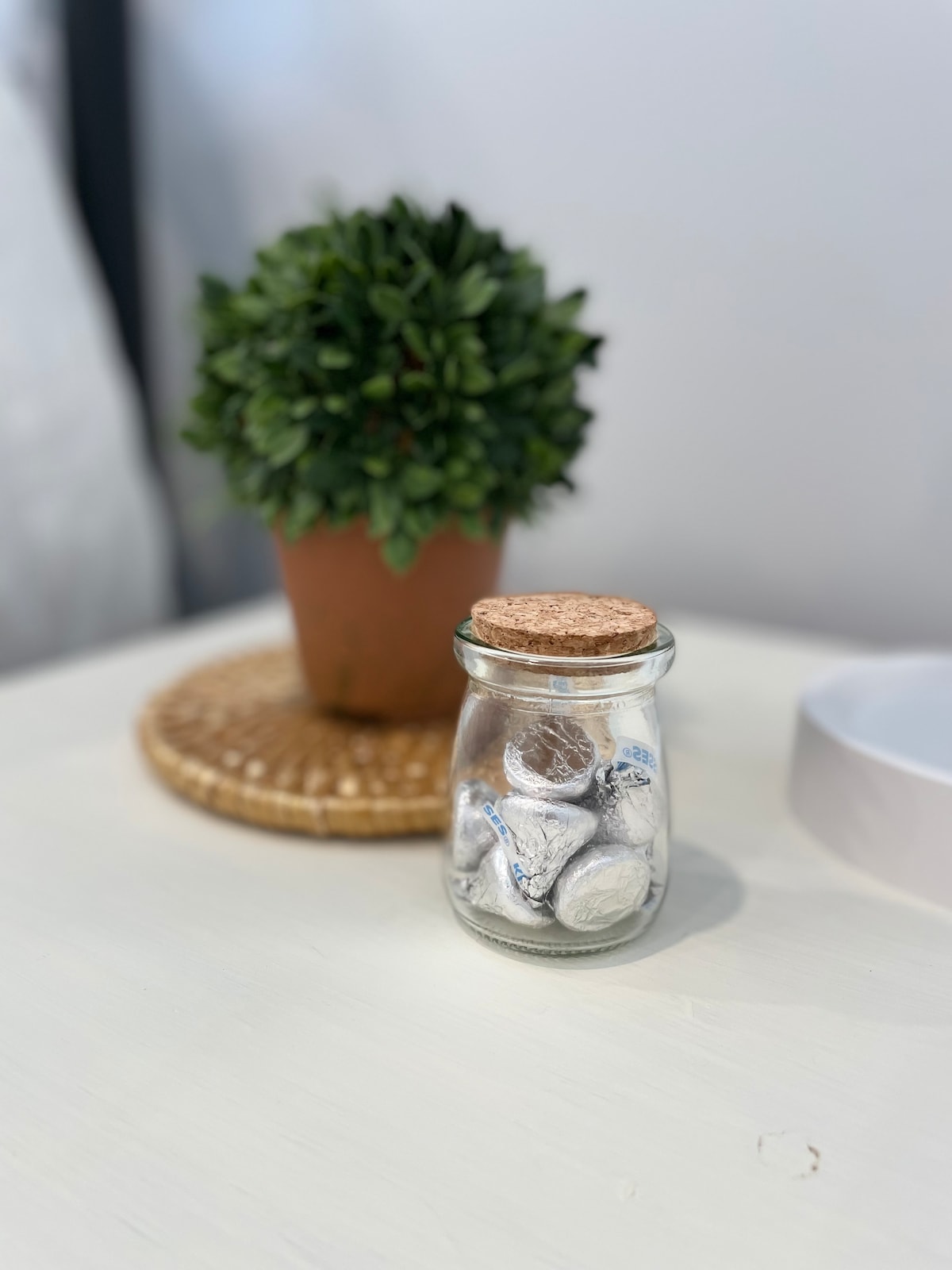 A small glass jar, topped with a cork lid, contains assorted wrapped candies. The jar is placed on a woven mat on a light-colored table. In the background, a potted plant adds a touch of greenery, complementing the serene setting.