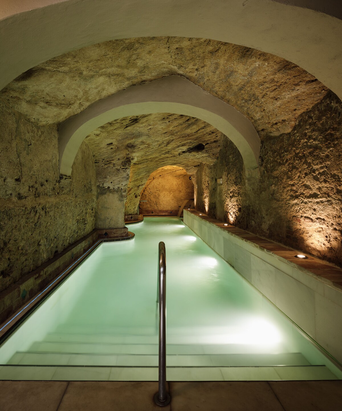 A serene indoor pool is featured within a cave-like setting, with stone walls illuminated by soft lighting. The calm water reflects the natural textures of the surroundings, while a railing guides the way along the edge of the pool.