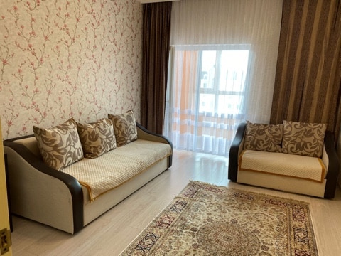 A living area is furnished with a spacious sofa and a matching armchair, both adorned with decorative cushions. A large window allows natural light to brighten the room, which features a light-colored wooden floor and a patterned area rug.