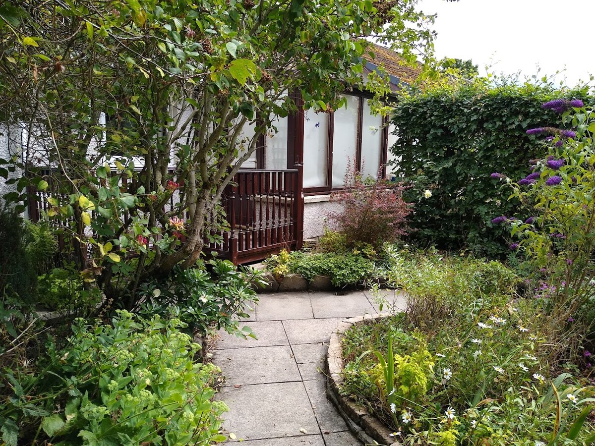 A well-maintained garden path winds through lush greenery, leading to the entrance of the cottage. The pathway is bordered by various plants and flowers, with vibrant foliage providing a natural backdrop. The conservatory features large windows that allow light into the inviting interior.