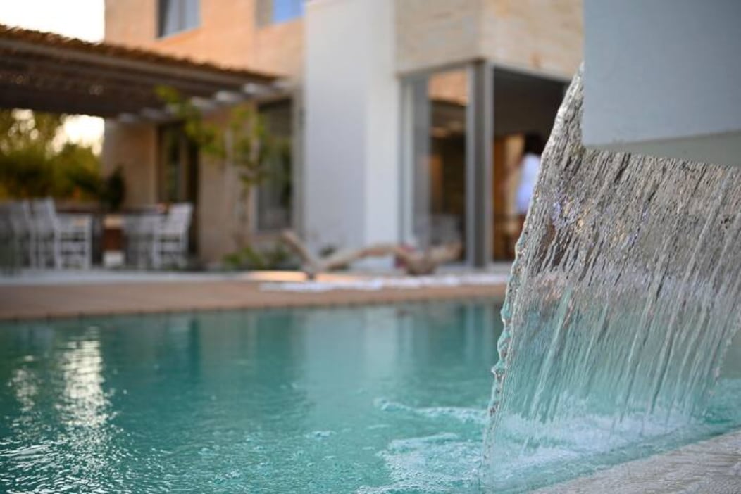Water cascades gently from a stone feature into a clear pool, creating a serene environment. In the background, a shaded pergola and outdoor dining area are visible, enhancing the relaxed atmosphere of the outdoor space surrounded by greenery.