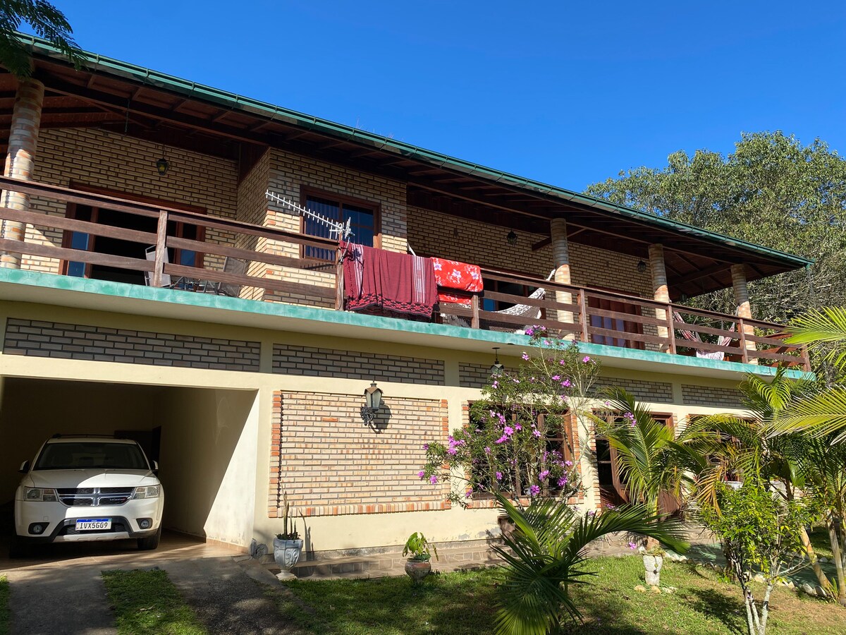 A spacious two-story house features a covered balcony with colorful blankets hanging to dry. The brick exterior is complemented by surrounding greenery and a driveway with a parked car. The entrance is framed by landscaping, including flowering plants.