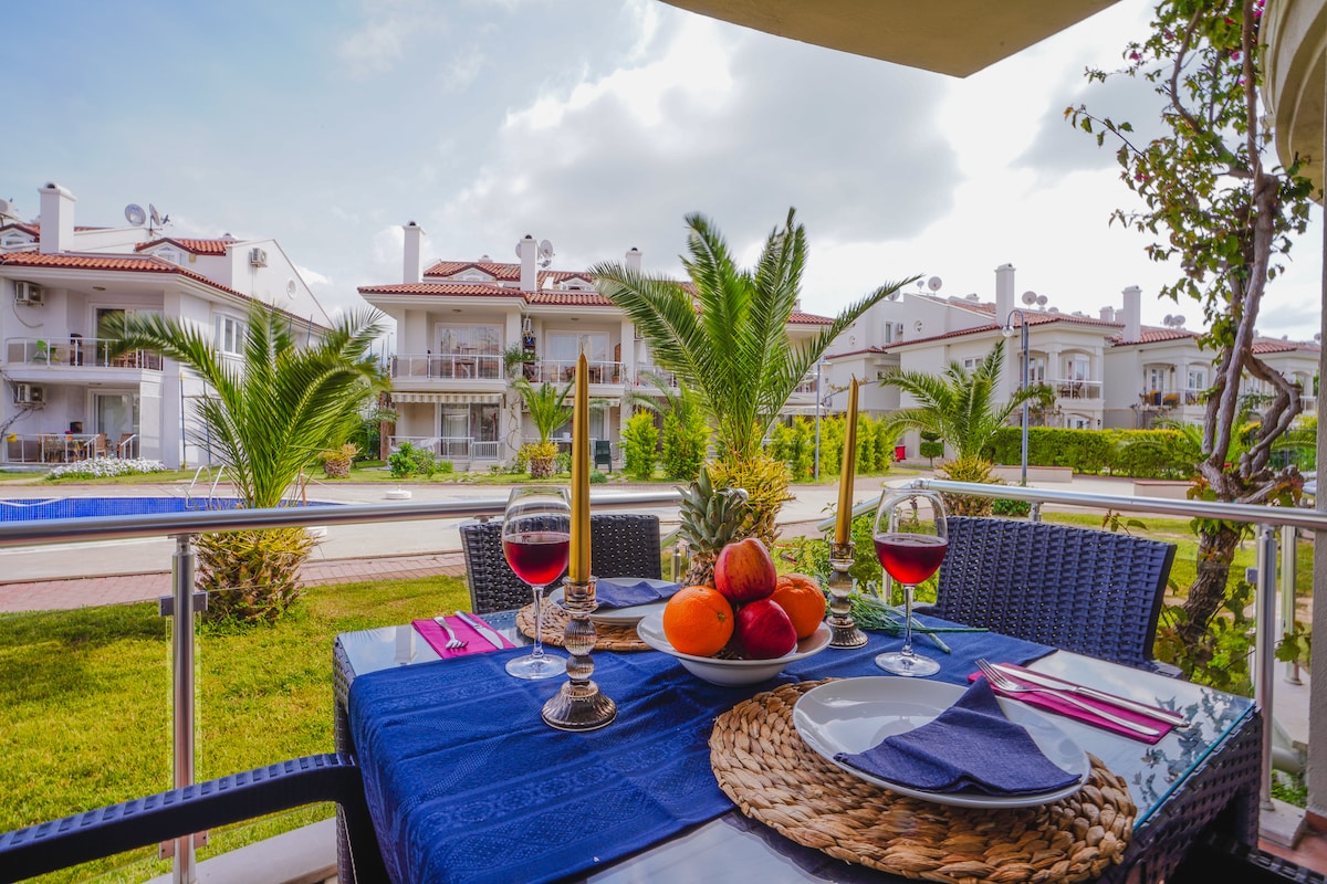 A dining table is set with plates and drinks on a balcony, surrounded by greenery and palm trees. A view of the neighboring buildings and pools can be seen in the background, creating a relaxed atmosphere for outdoor dining.