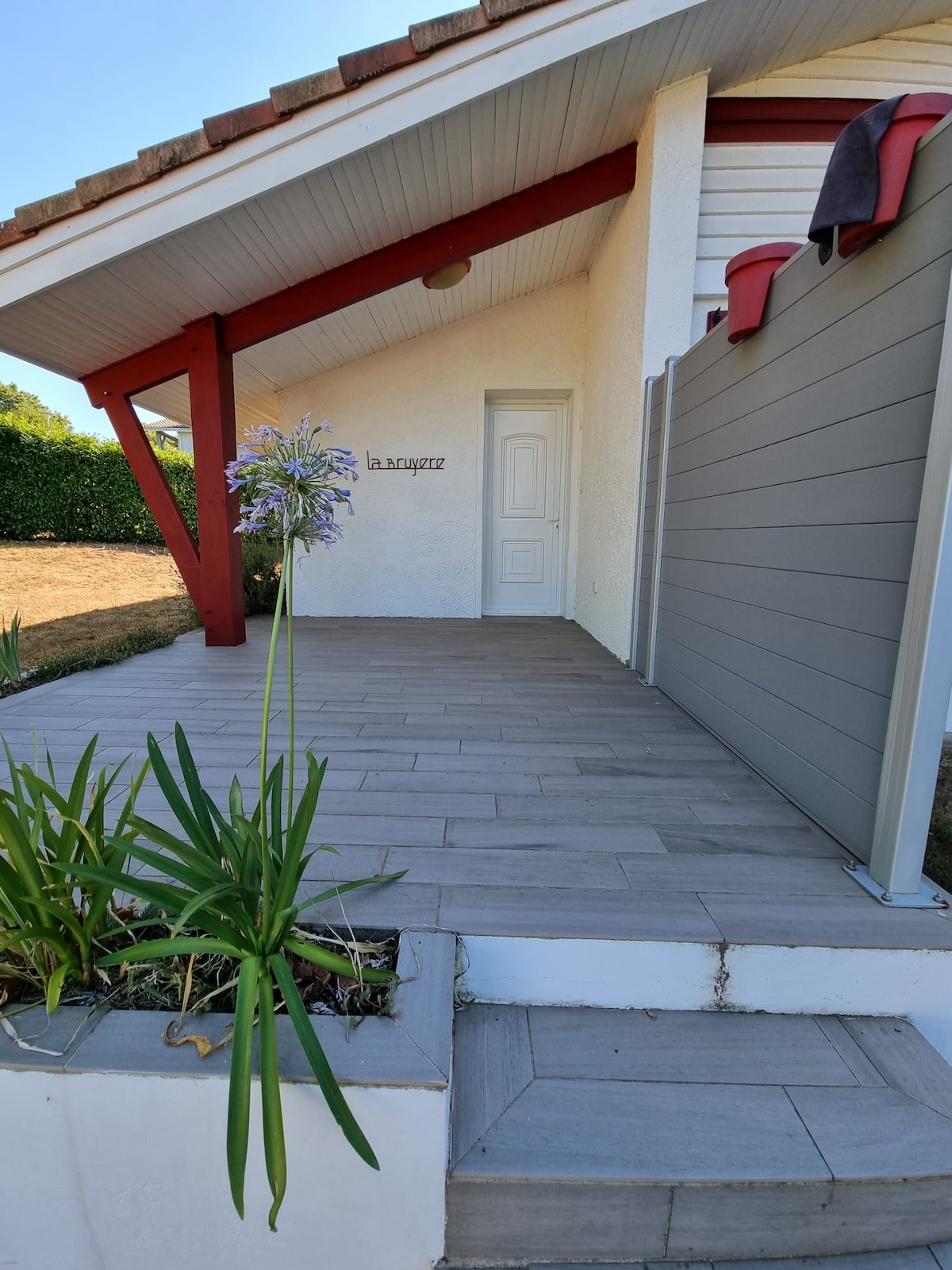 A front porch is displayed, featuring a covered area with wooden beams and a contemporary grey fence. A planter with green foliage and purple flowers adds a touch of nature, while the entrance door blends seamlessly into the white exterior wall.
