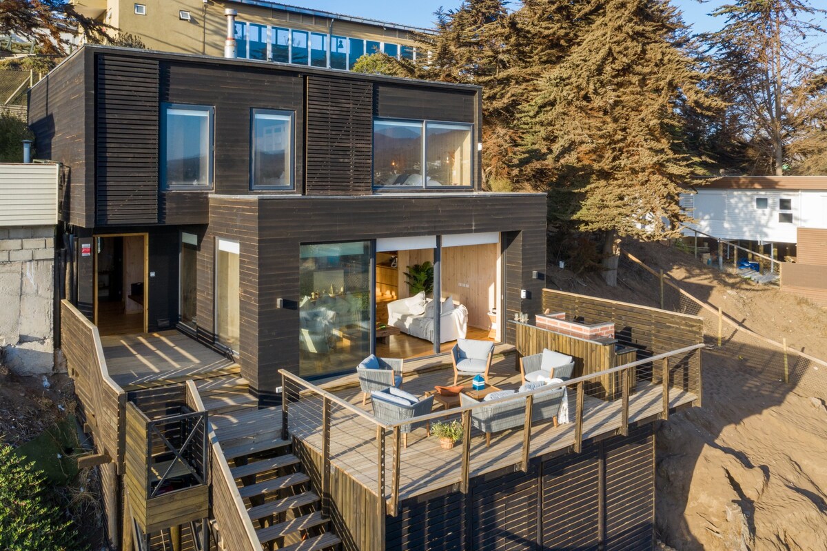 An exterior view of a modern two-story house is shown, featuring large windows and a blend of wood and glass. A spacious deck surrounds the home, furnished with seating areas and a barbecue space, while the sloping landscape is adorned with greenery.