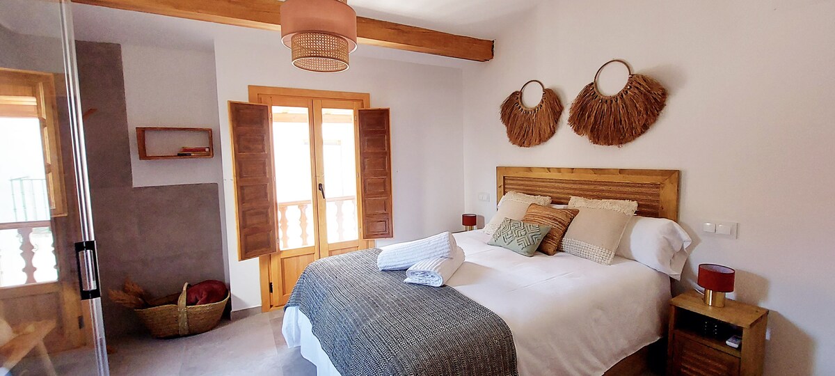 A brightly lit bedroom features a spacious bed adorned with neutral linens and decorative pillows. Two woven wall hangings add texture, while light wood accents complement the warm atmosphere. The large windows, flanked by wooden shutters, offer natural light, and a cozy basket is placed nearby.