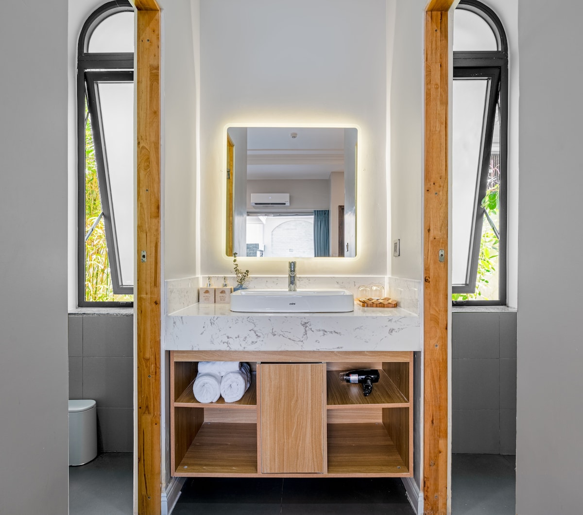 A modern bathroom features a sleek countertop with a well-lit mirror above a spacious sink. Wooden cabinetry provides storage, showcasing neatly rolled towels. Natural light enters through two framed windows, enhancing the airy feel of the space.