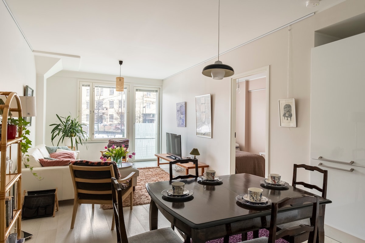 The open living space is illuminated by natural light from large windows. A dining table is set for four, with decorative cups. A comfortable seating area is positioned nearby, featuring a plush sofa and TV. A well-defined entry leads to the private sleeping area.