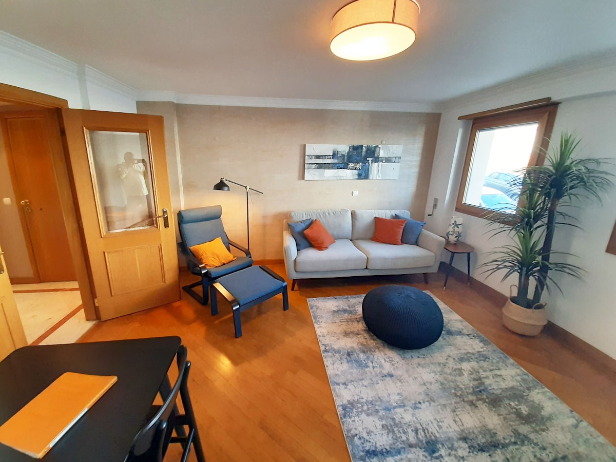 The living area features a light-colored sofa with orange accent cushions, paired with a dark blue chair. A modern floor lamp provides soft lighting. A large area rug in muted tones adds warmth, and a potted plant enhances the inviting ambiance.