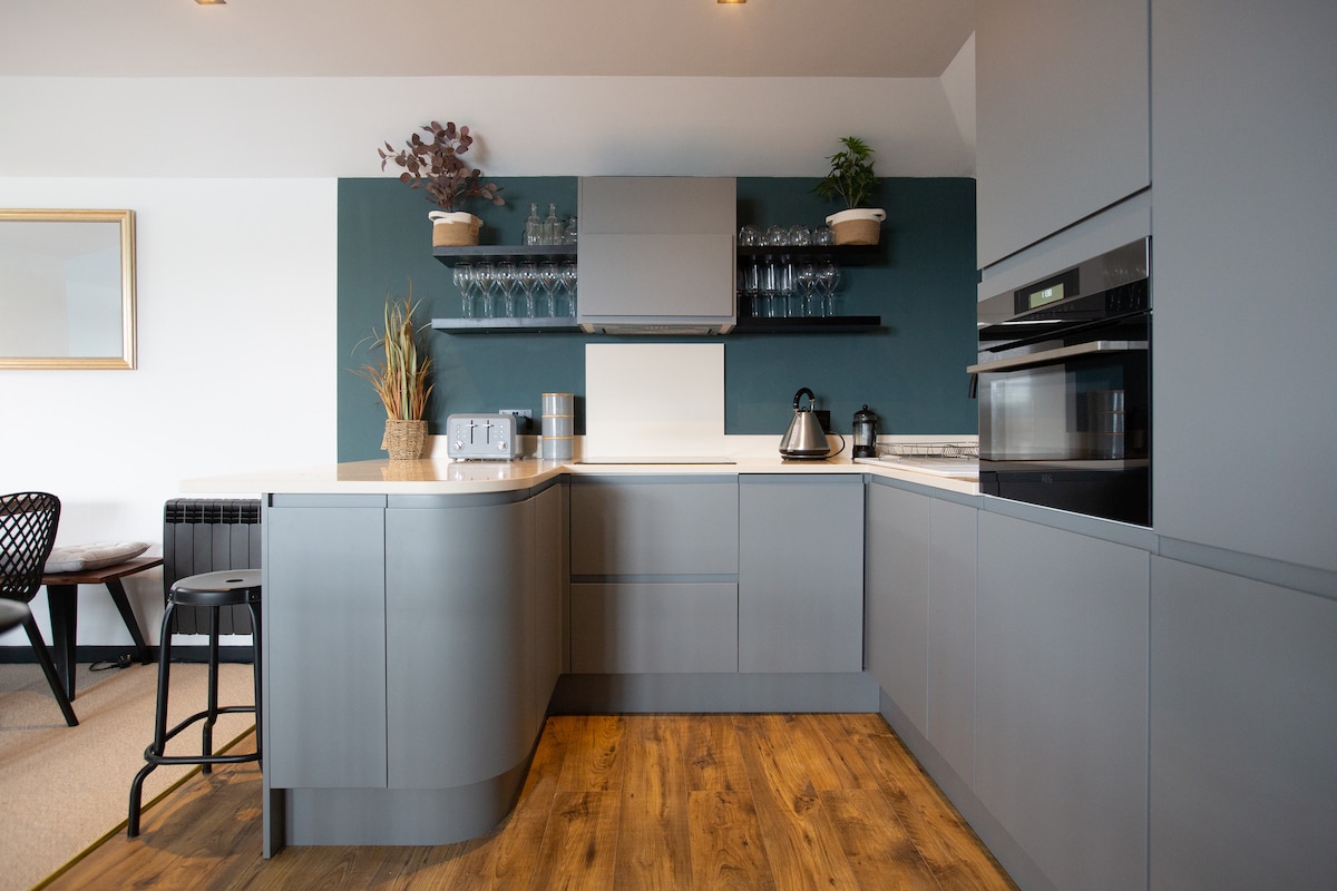 A modern kitchen features grey cabinetry with a sleek countertop, complemented by an arrangement of glasses on shelves above. A kettle and toaster are prominently displayed. Adjacent seating is seen, along with minimal decor of plants and wall art adding subtle elegance.