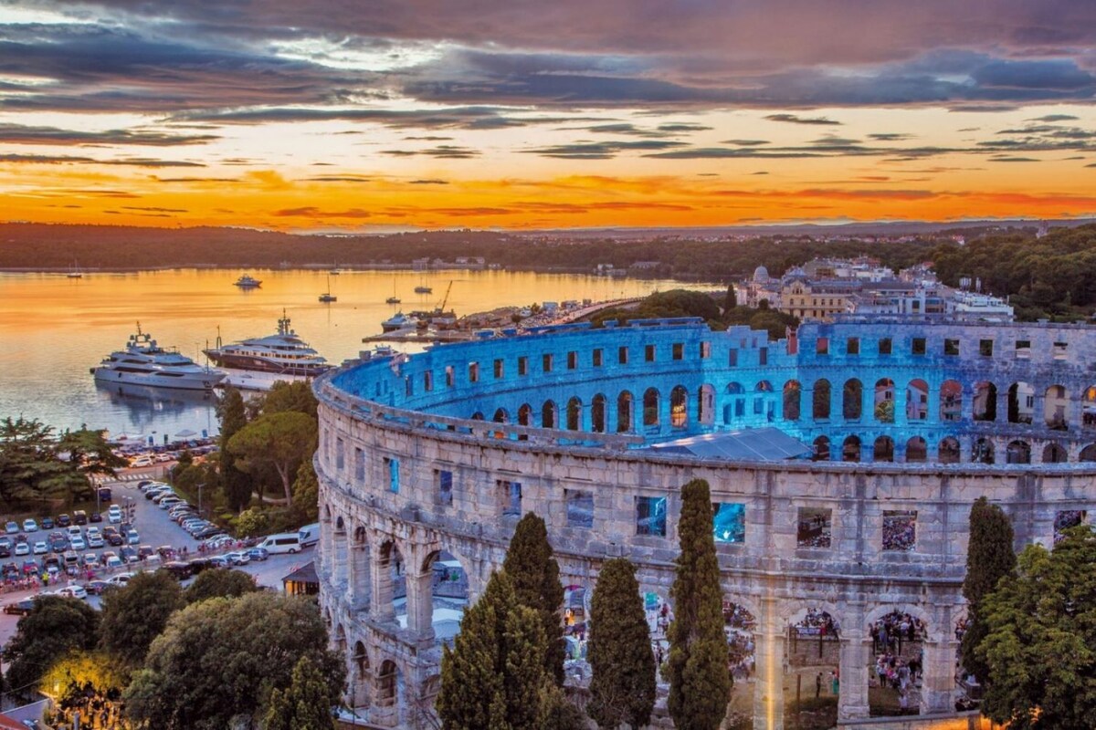 The image captures the historic amphitheater amidst a scenic sunset. The sky transitions through warm hues, while yachts are seen docked in the water. Lush greenery and cityscapes surround the ancient structure, creating a peaceful coastal atmosphere.