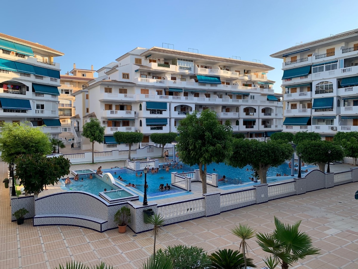 A spacious outdoor area features a large swimming pool surrounded by tropical trees and well-maintained patios. The surrounding buildings display balconies with awnings. Residents and guests can be seen enjoying the pool, contributing to a lively communal atmosphere.