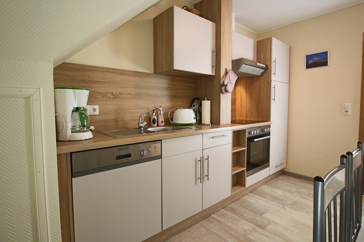 The kitchen features modern cabinetry with a wood finish in warm tones. Stainless steel appliances, including a dishwasher and oven, are included. Counter space is available for meal preparation, complemented by a coffee maker and kettle for convenience.