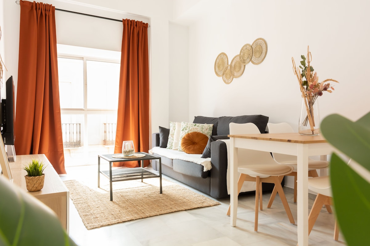 A bright living area features a dark sofa adorned with decorative cushions. A simple wooden table and light-colored chairs are positioned nearby. The space is complemented by elegant wall decor and large windows, which are framed by flowing orange curtains, allowing natural light to fill the room.