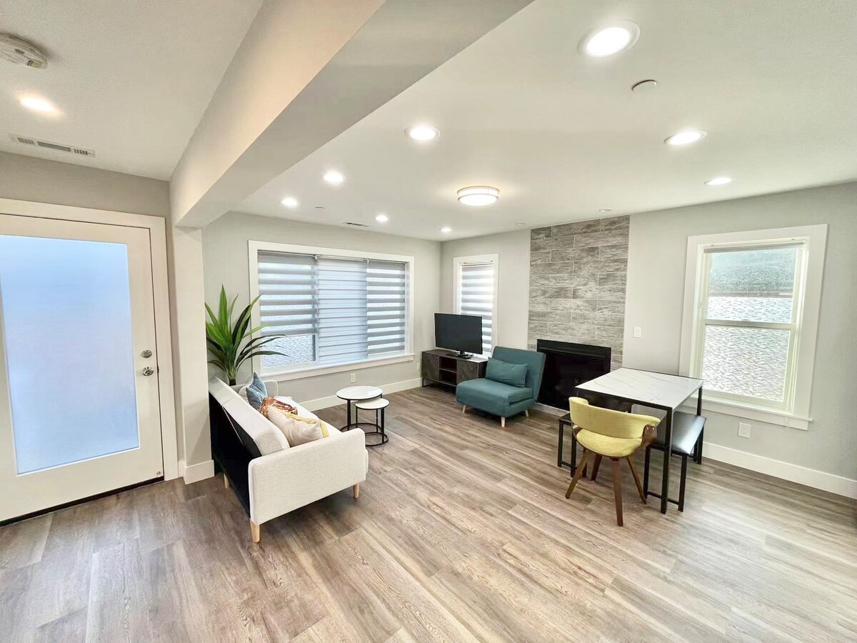 A modern living area is presented with sleek hardwood flooring and neutral-colored walls. A comfortable sofa and armchair are arranged around a coffee table, while a television stands against a stone accent wall. Natural light filters through lined windows, creating an inviting ambiance.