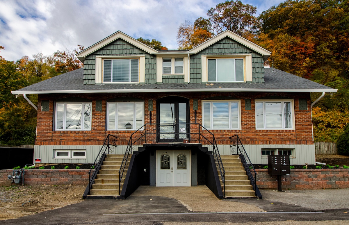The exterior of the property showcases a two-story brick building with a gabled roof and green shingles. Symmetrical windows are positioned on the upper and lower levels. A central entrance is flanked by two sets of stairs, leading to a landing with decorative railings.