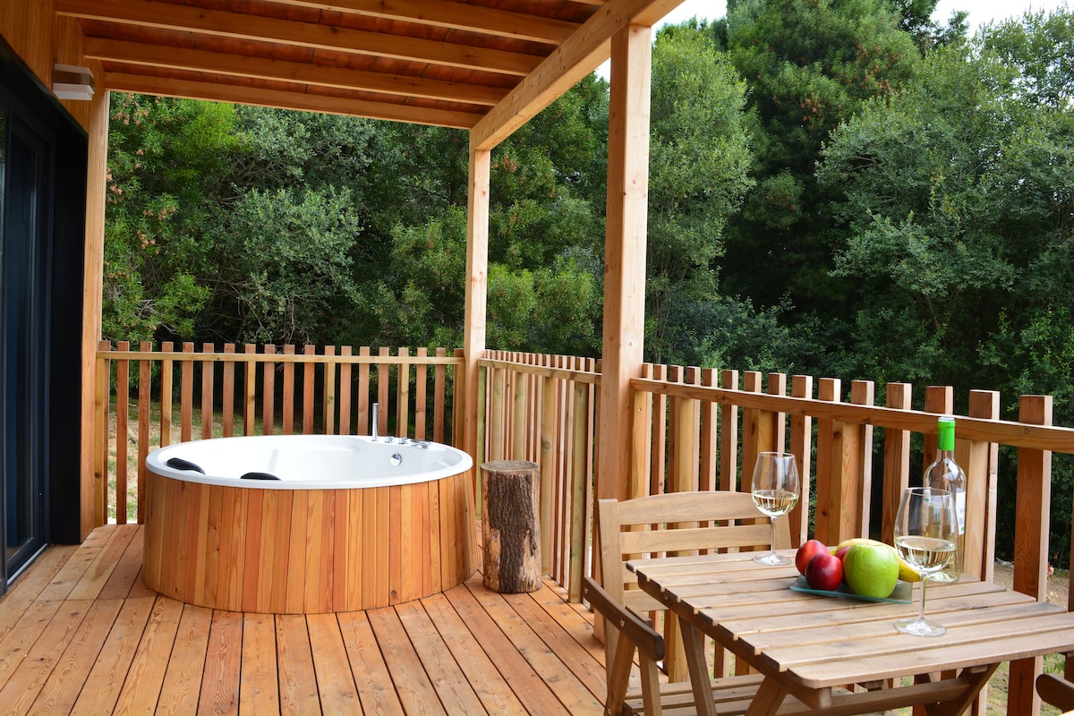 A wooden terrace is shown, featuring a round jacuzzi surrounded by a natural wooden railing. A small table holds three apples and a bottle, with two glasses nearby. Lush greenery can be seen in the background, enhancing the serene atmosphere.
