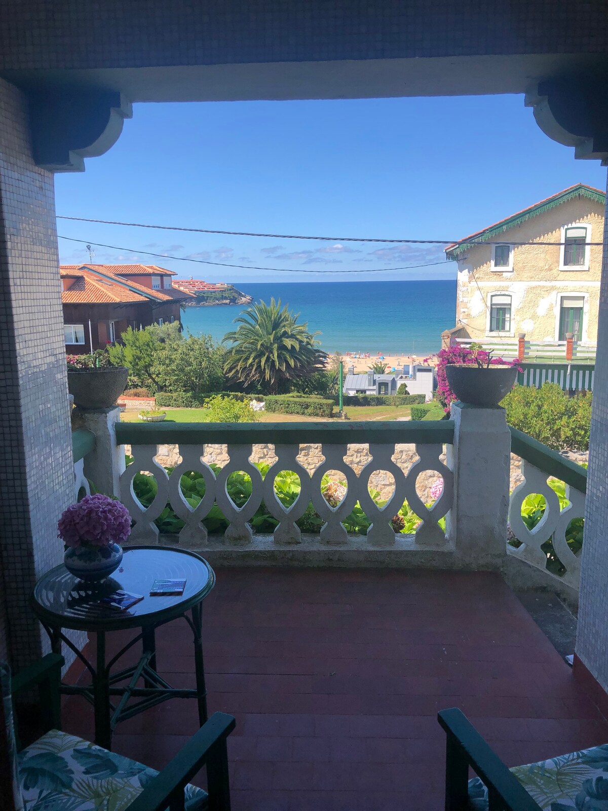 A view from a terrace showcases the ocean and sandy beach, framed by a decorative railing. Potted plants add a touch of greenery, while nearby buildings, including a charming house, complete the scene. The sky is clear, highlighting the serene coastal atmosphere.