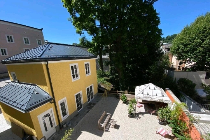 A view of a charming yellow building surrounded by greenery captures a serene outdoor space. A white canopy is positioned over a seating area, complemented by a rustic bench. Bright sunlight illuminates the scene, creating a welcoming atmosphere for relaxation.