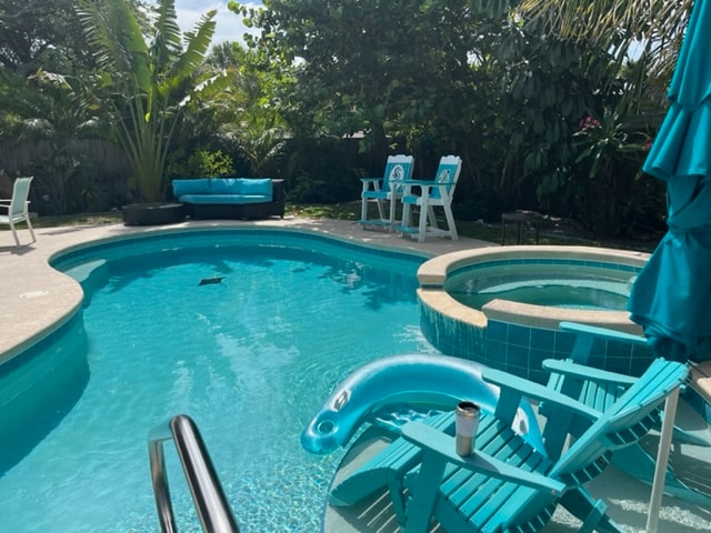 The image features a private heated pool surrounded by lush tropical landscaping, including palm trees and other greenery. A hot tub is integrated into the pool area. Bright turquoise lounge chairs and a small table are strategically placed near the pool.
