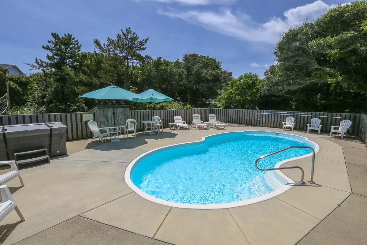 A private pool area is surrounded by several lounge chairs and shaded umbrellas. The pool features a curvy shape, with a smooth deck providing ample space for relaxation. Lush trees provide a natural backdrop, enhancing the sense of seclusion.