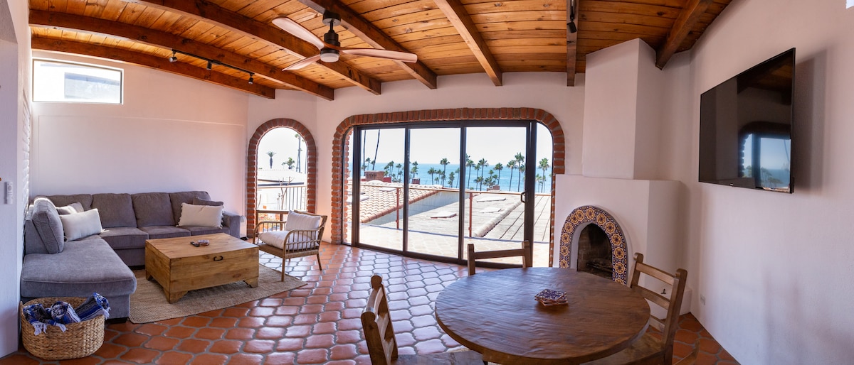 A well-lit living area features a spacious gray sofa and a rustic wooden coffee table. Large glass doors provide a panoramic ocean view, framed by tropical palm trees. The room includes a round table with chairs, with traditional tilework surrounding an alcove fireplace.
